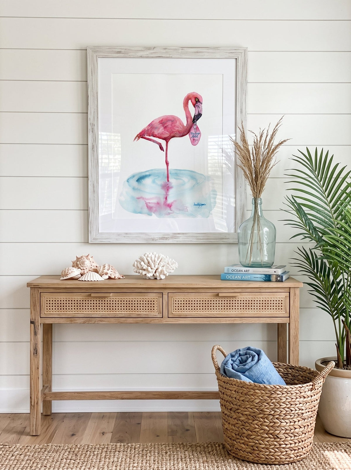 A watercolor painting titled "Flamingo Motel davidkjonesart.jpg", featuring a pink flamingo standing on one leg in a shallow pool of light blue water with its pink reflection visible below. The flamingo is holding a retro pink motel keychain in its beak with the text "Flamingo Motel Room 7". The artwork is signed by the artist in the bottom right corner.