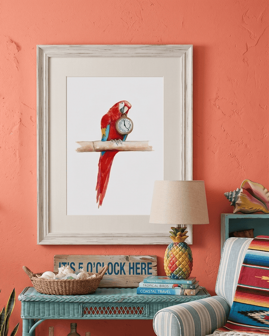 Framed watercolor painting by artist David K. Jones titled "It's Five O'clock HERE," featuring a vibrant scarlet macaw parrot perched on a bamboo branch against a clean white background. The red, blue, and yellow parrot holds a vintage pocket watch in its beak, with the hands explicitly pointing exactly to 5:00. Hung on a coral colored wall over a turquoise wicker table with tropical decor.
