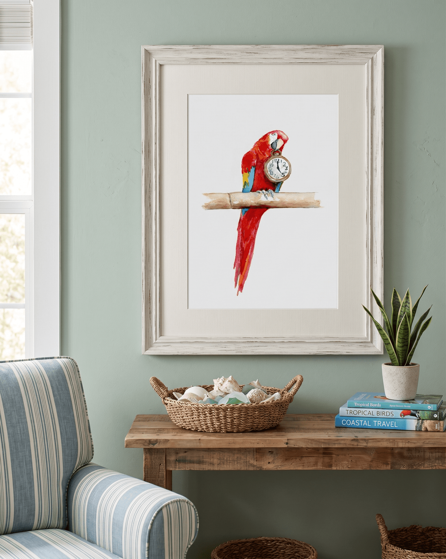 Framed watercolor painting by artist David K. Jones titled "It's Five O'clock HERE," featuring a vibrant scarlet macaw parrot perched on a bamboo branch against a clean white background. The red, blue, and yellow parrot holds a vintage pocket watch in its beak, with the hands explicitly pointing exactly to 5:00. Hung on a light green wall over a table with seashells.