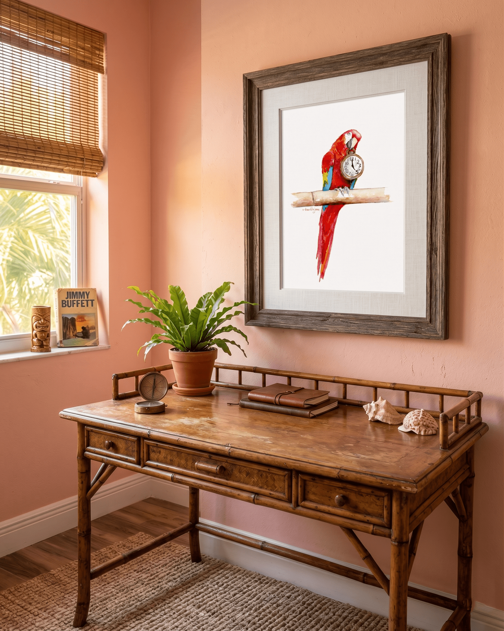 Framed watercolor painting by artist David K. Jones titled "It's Five O'clock HERE," featuring a vibrant scarlet macaw parrot perched on a bamboo branch against a clean white background. The red, blue, and yellow parrot holds a vintage pocket watch in its beak, with the hands explicitly pointing exactly to 5:00. Hung on a coral colored wall over a desk next to a window showing a palm tree.