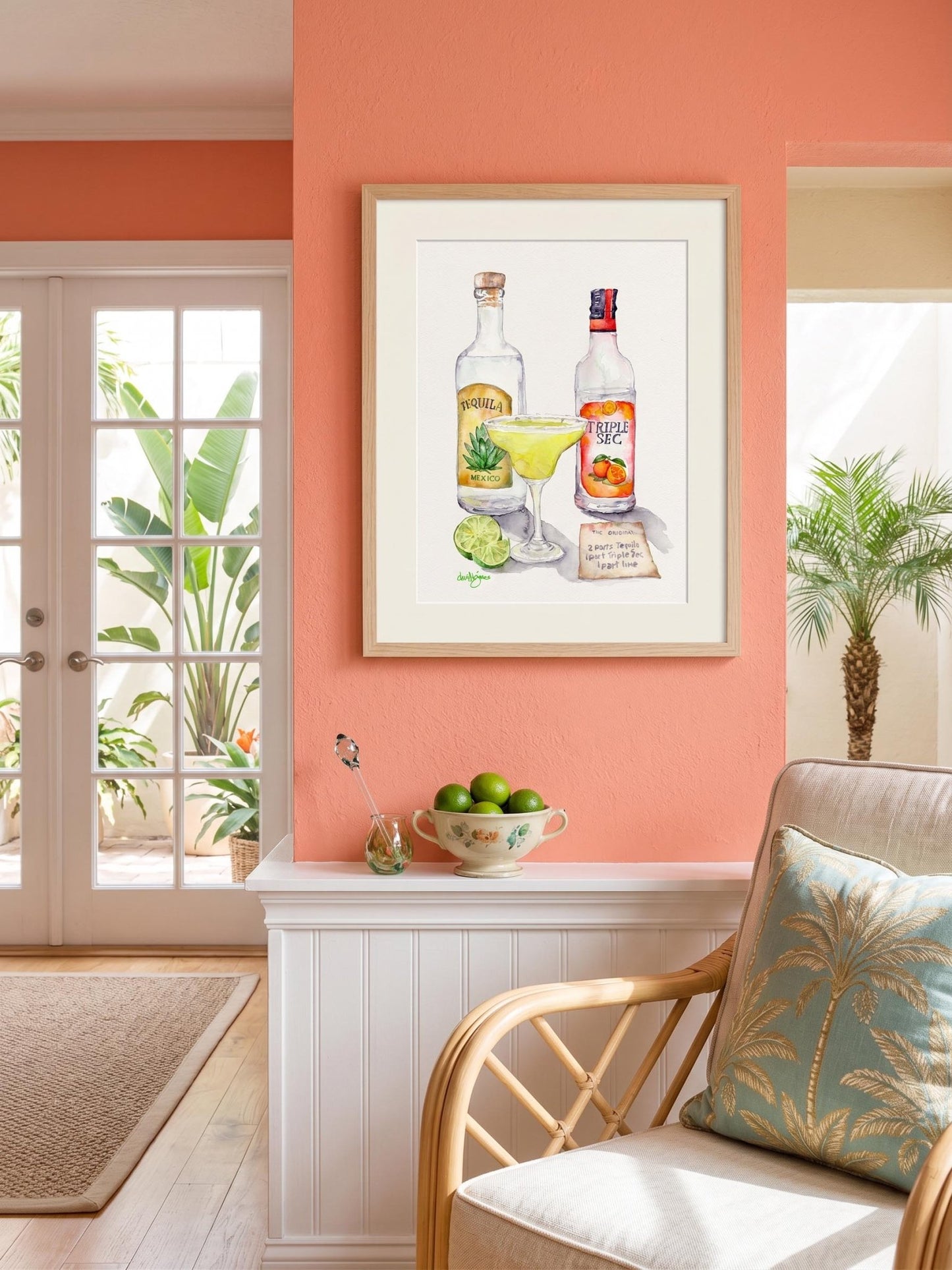 Loose and colorful watercolor print of a margarita cocktail with tequila and Triple Sec bottles. The artwork includes a margarita glass, lime slices, and a recipe card. The print is framed and displayed on a coral-colored wall in a Kewy West style room. Art by David K. Jones