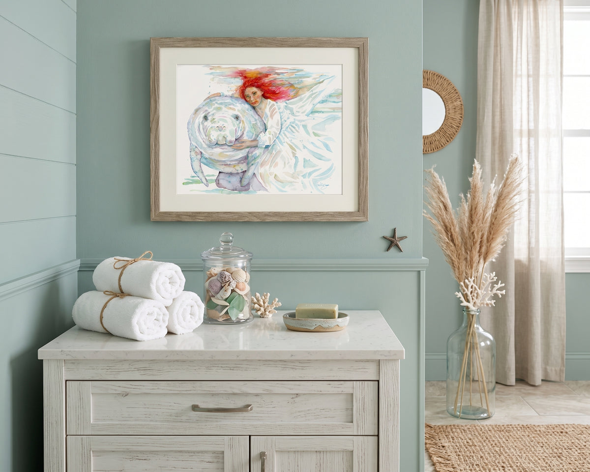 "Mermaids" watercolor painting by David K. Jones, depicting a woman with flowing red hair in a white dress gently embracing a manatee underwater. styled in a coastal-themed room above a white dresser.