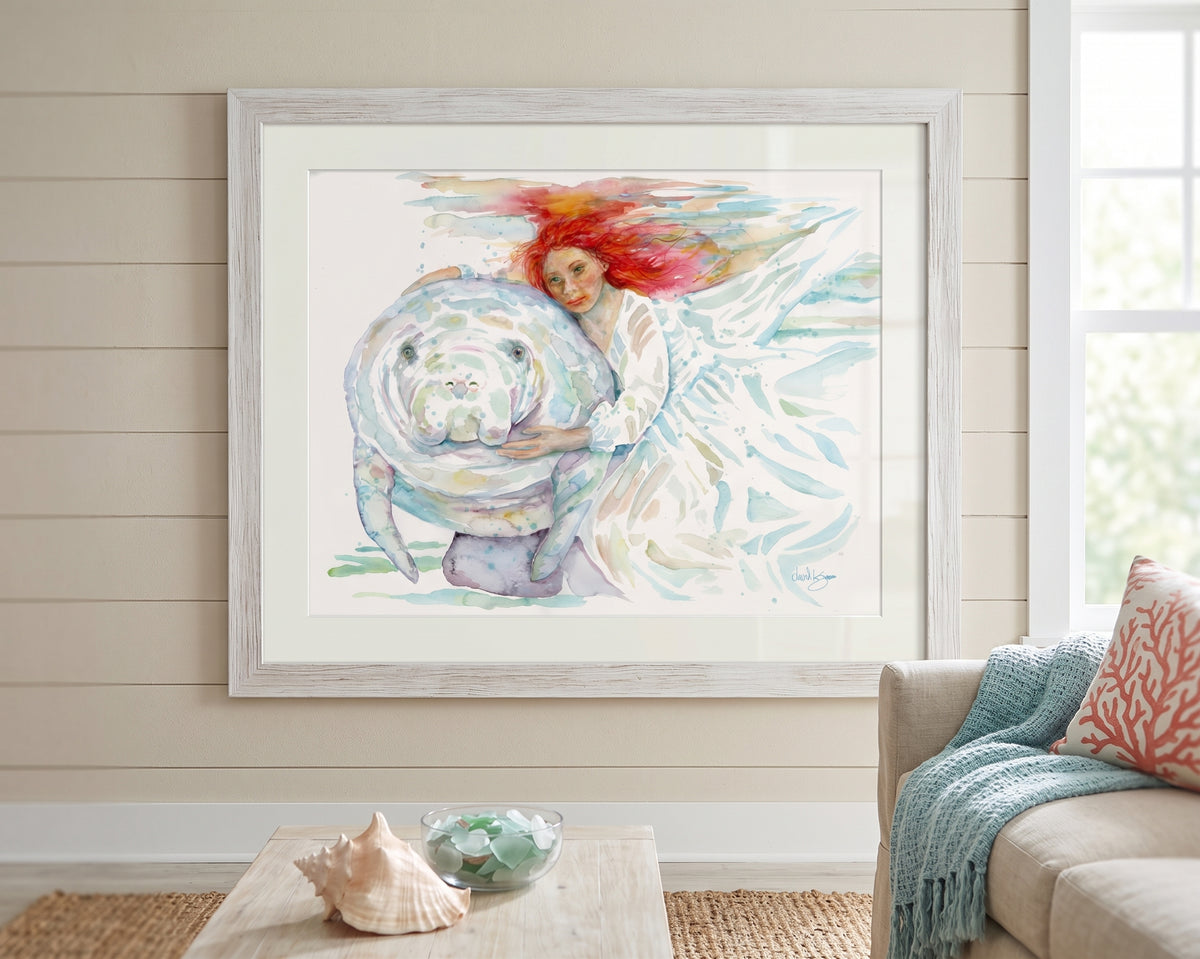 "Mermaids" watercolor painting by David K. Jones, depicting a woman with flowing red hair in a white dress gently embracing a manatee underwater., displayed on a beige shiplap wall above a coffee table in a styled living room.