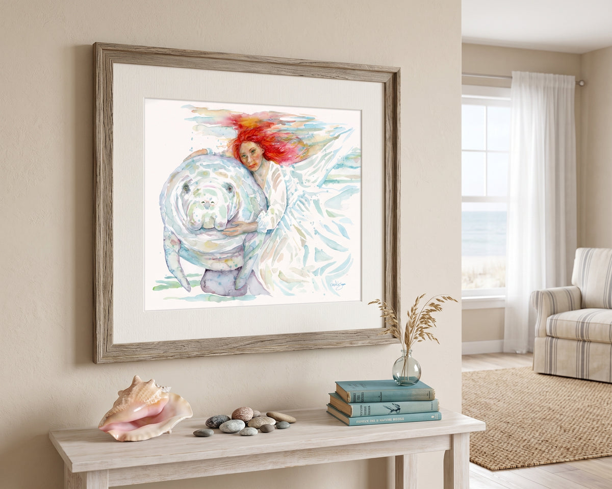 "Mermaids" watercolor painting by David K. Jones, depicting a woman with flowing red hair in a white dress gently embracing a manatee underwater., displayed in a rustic wood frame on a beige wall above a console table in a coastal-themed living room.