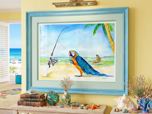 A framed print of David K. Jones's tropical watercolor "Parrothead Fish Tale" displayed in a beach-themed room. The painting is set in a wide, light blue wooden frame with a seafoam green mat, hanging on a soft yellow wall. Below the art, a wooden console table is styled with coastal decor, including sea glass floats, seashells, a starfish, coral, and vintage books. A doorway opens to a sunny ocean beach view.