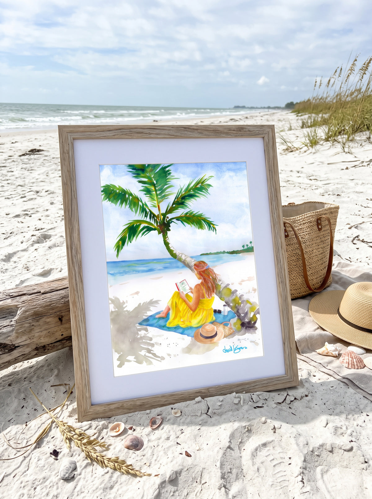 
 "Turning Pages in Paradise" by David K. Jones. A watercolor painting depicting a woman reading a book on a tropical beach. She wears a bright yellow dress and sits on a blue towel beneath a curved palm tree with green fronds. A straw hat with a black band, sunglasses, a tumbler, and a woven tote bag rest on the white sand nearby, with blue oceA framed beach art print displayed on a sandy beach. The print shows a woman in a yellow dress sitting under a palm tree by the ocean.
