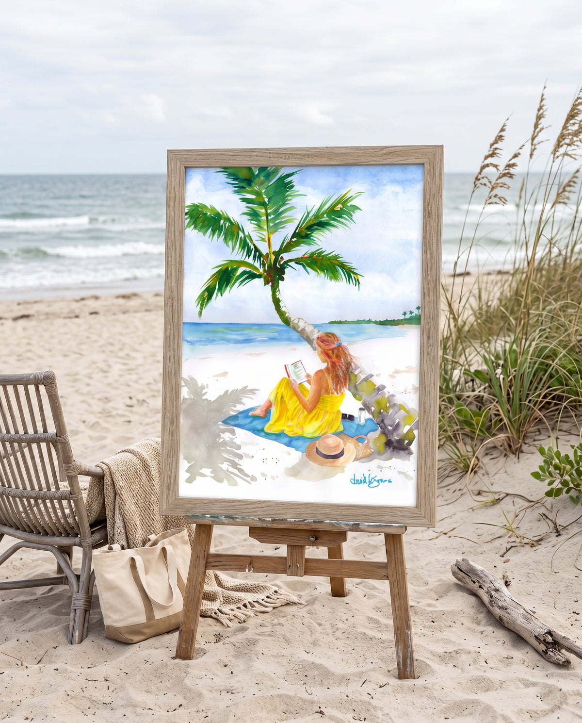 A framed beach art print displayed on a wooden easel at the beach. "Turning Pages in Paradise" by David K. Jones. A watercolor painting depicting a woman reading a book on a tropical beach. She wears a bright yellow dress and sits on a blue towel beneath a curved palm tree with green fronds. A straw hat with a black band, sunglasses, a tumbler, and a woven tote bag rest on the white sand nearby, with blue ocean water in the background.