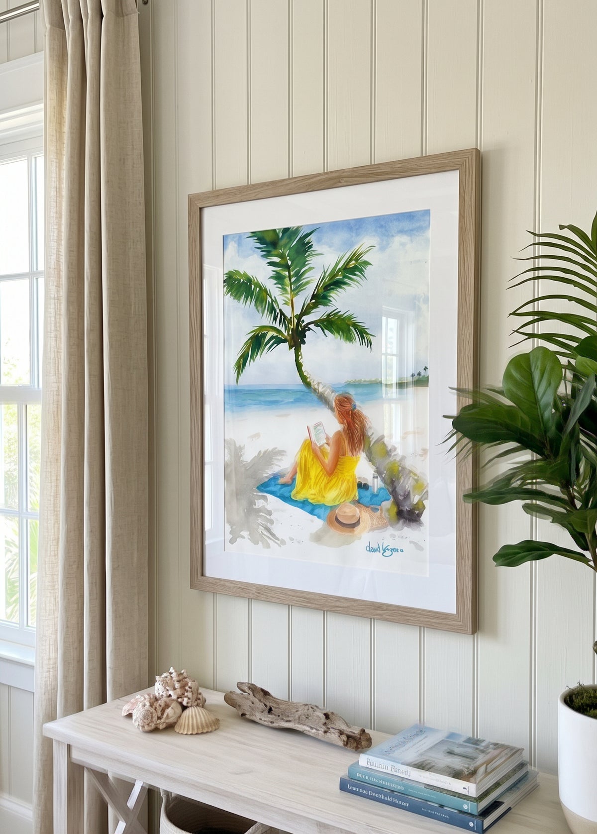 A framed beach art print hangs on a cream paneled wall above a console table. "Turning Pages in Paradise" by David K. Jones. A watercolor painting depicting a woman reading a book on a tropical beach. She wears a bright yellow dress and sits on a blue towel beneath a curved palm tree with green fronds. A straw hat with a black band, sunglasses, a tumbler, and a woven tote bag rest on the white sand nearby, with blue ocean water in the background.