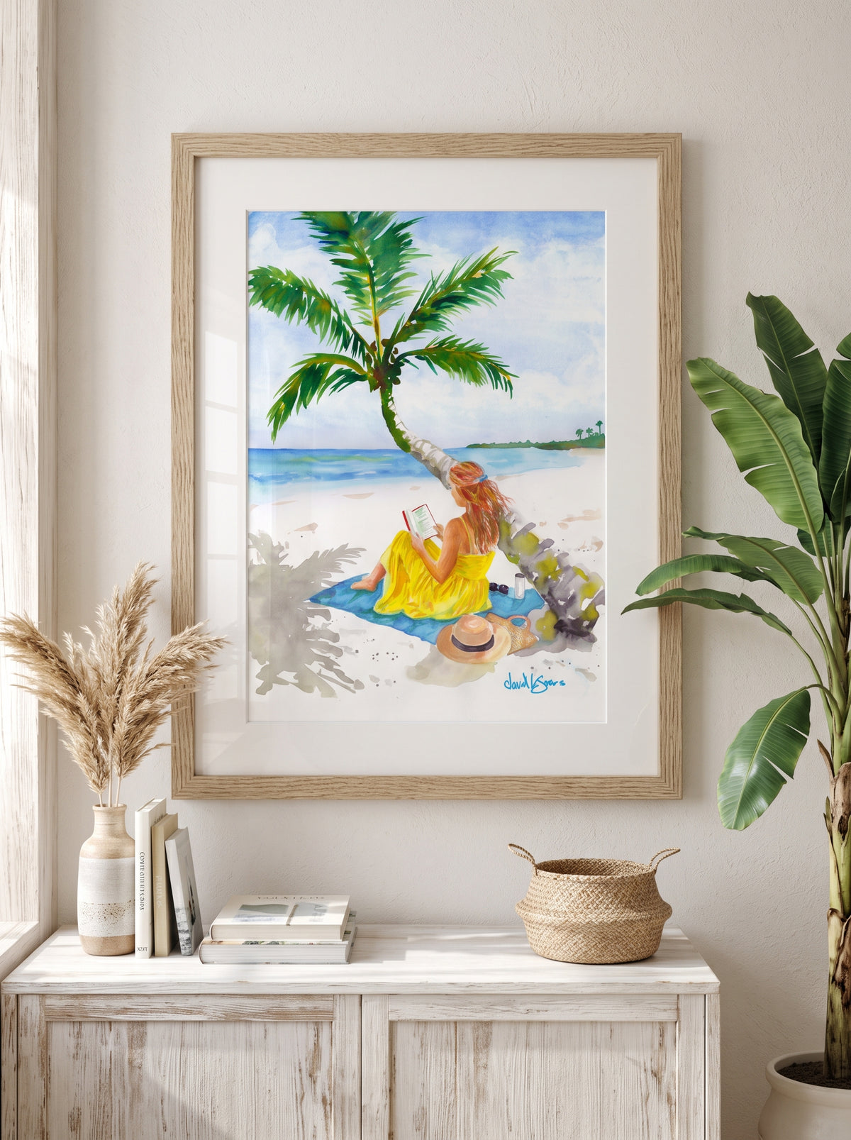 "Turning Pages in Paradise" by David K. Jones. A watercolor painting depicting a woman reading a book on a tropical beach. She wears a bright yellow dress and sits on a blue towel beneath a curved palm tree with green fronds. A straw hat with a black band, sunglasses, a tumbler, and a woven tote bag rest on the white sand nearby, with blue ocean water in the background. displayed on a white console table in a styled room with a potted plant and decorative items.