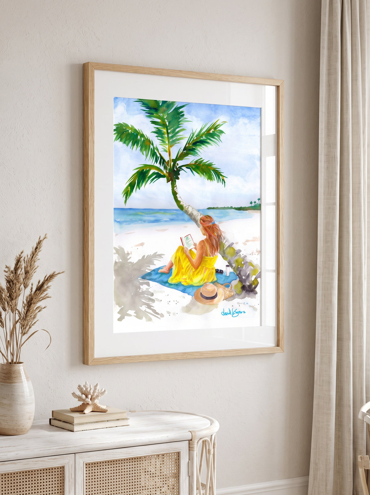 "Turning Pages in Paradise" by David K. Jones. A watercolor painting depicting a woman reading a book on a tropical beach. She wears a bright yellow dress and sits on a blue towel beneath a curved palm tree with green fronds. A straw hat with a black band, sunglasses, a tumbler, and a woven tote bag rest on the white sand nearby, with blue ocean water in the background. hung on a light beige wall above a white console table with decorative items.