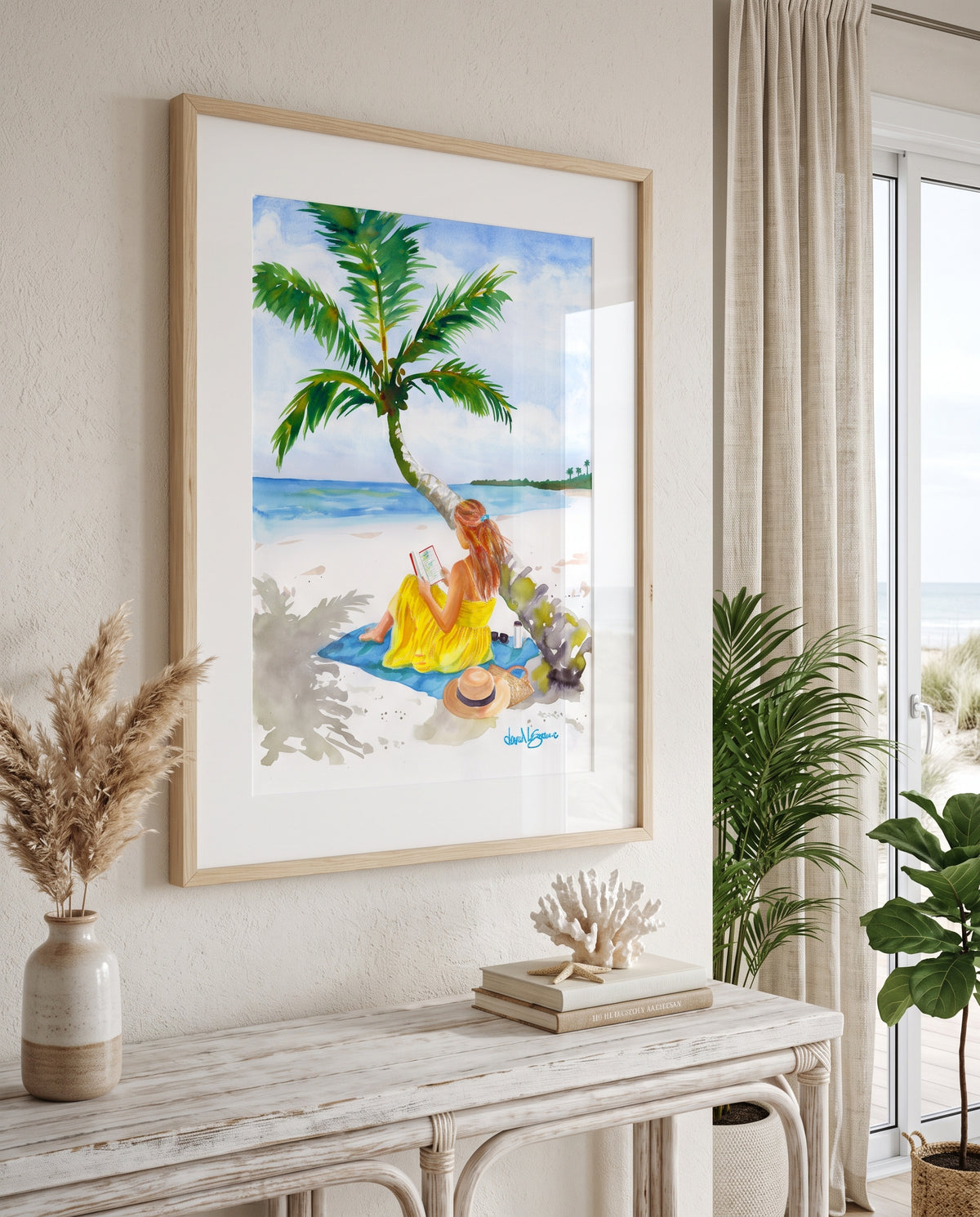 "Turning Pages in Paradise" by David K. Jones. A watercolor painting depicting a woman reading a book on a tropical beach. She wears a bright yellow dress and sits on a blue towel beneath a curved palm tree with green fronds. A straw hat with a black band, sunglasses, a tumbler, and a woven tote bag rest on the white sand nearby, with blue ocean water in the background.styled on a console table in a bright, modern room.